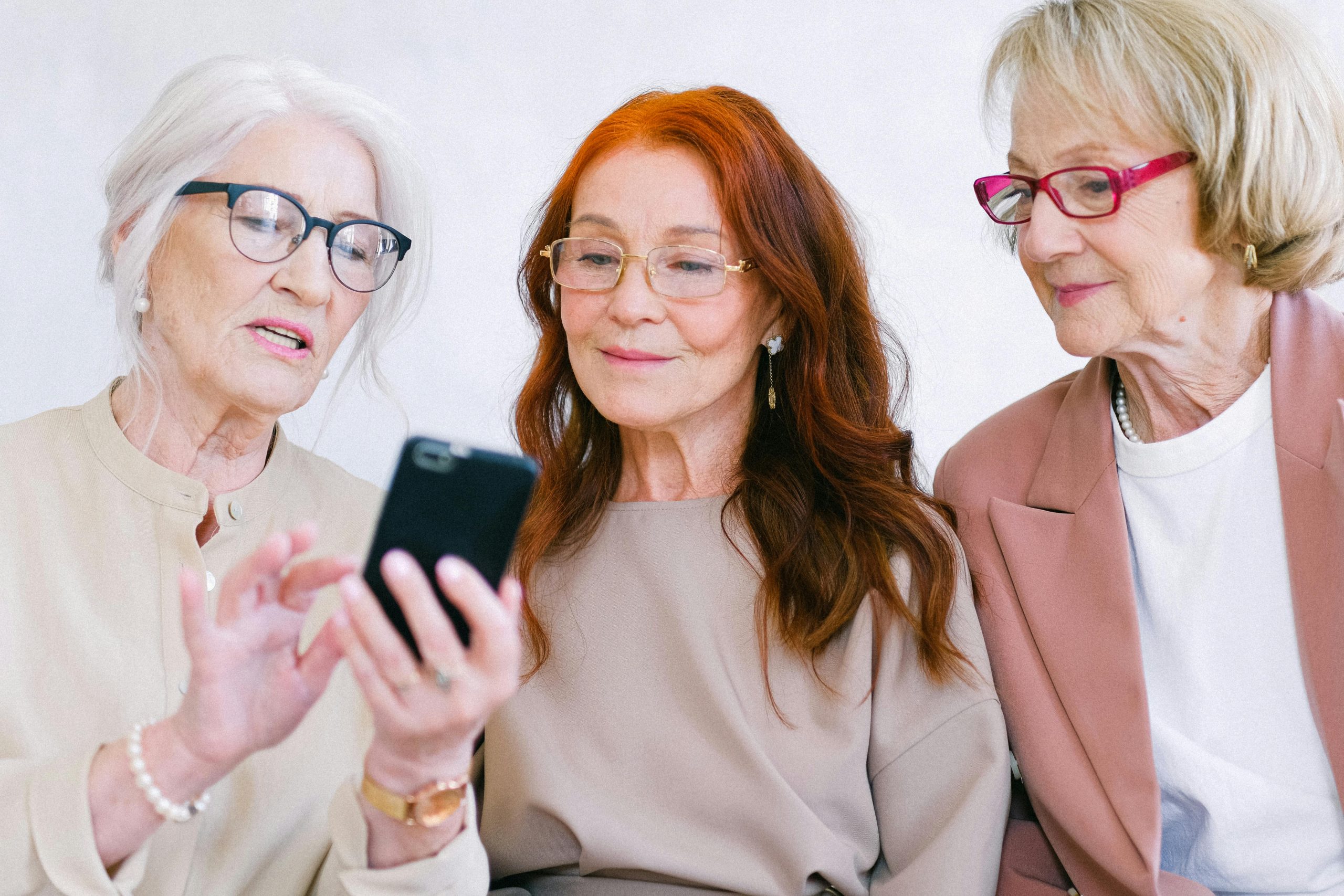 Three senior women enjoying a friendly moment together, sharing a smartphone and smiling.