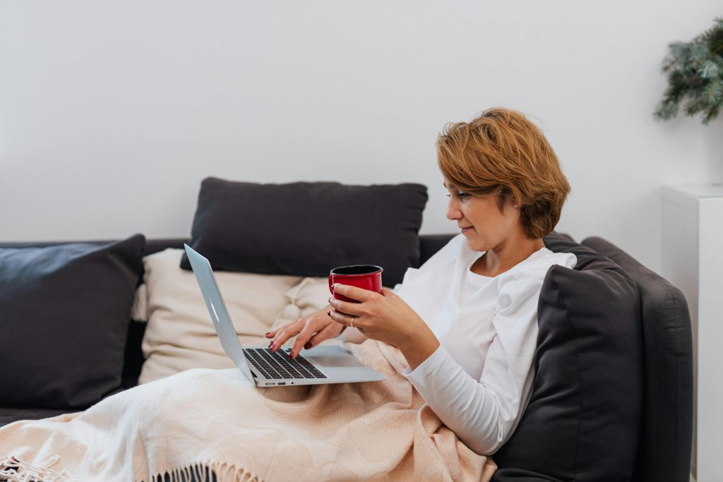 Adult woman enjoys coffee while working on a laptop in a cozy indoor setting.